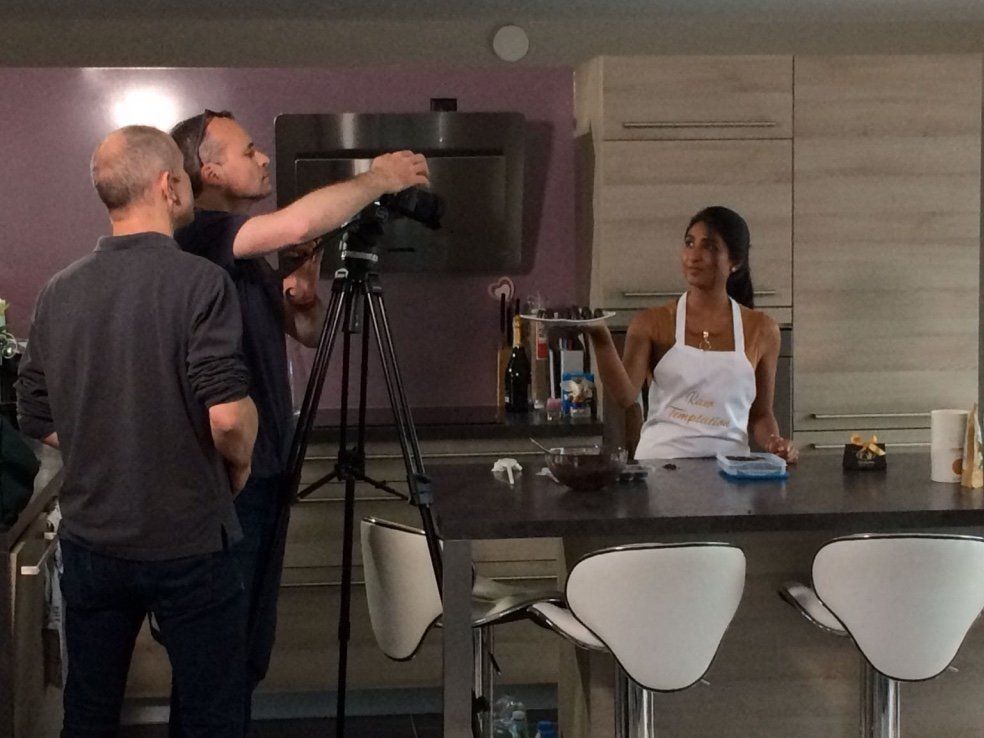 Candice Chetty in a Raw Temptation apron presenting a plate of pralines to a film crew in a kitchen