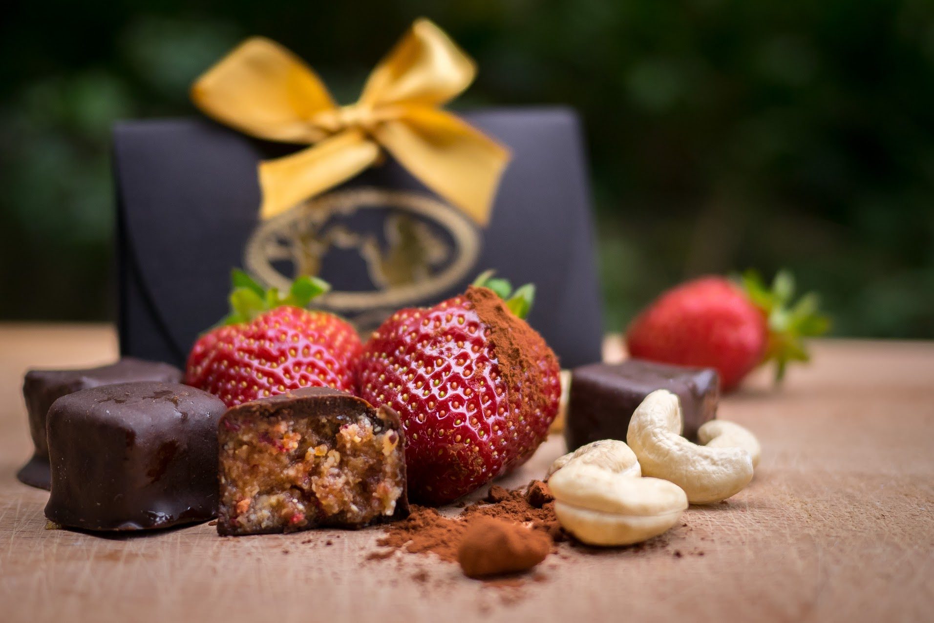 A black Raw Temptation gift box tied with gold ribbon, beside strawberry pralines, fresh strawberries and cashews